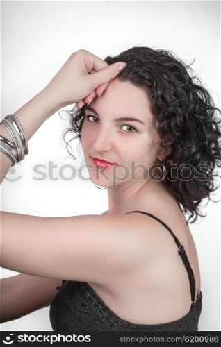 Portrait of a young woman with black hair and a black open shoulder top sitting sideways in front of a white background while looking towards the camera.