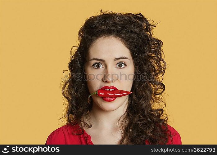 Portrait of a young woman with red chili pepper in mouth over colored background