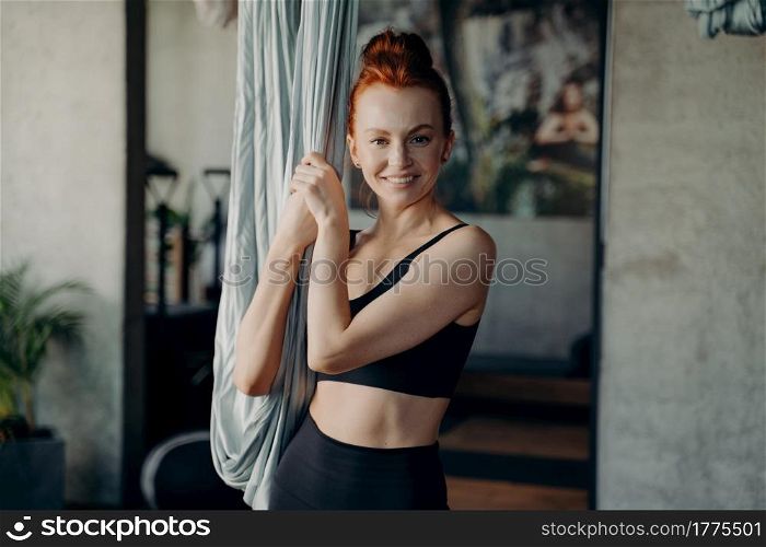 Portrait of active and healthy ginger woman looking happily at camera while standing by hammock, waiting for antigravity yoga class against blurred fitness studio background. Sport and fitness concept. Positive ginger girl standing near hammock smiling on camera waiting for aerial yoga class