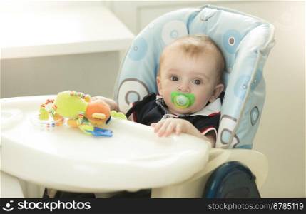 Portrait of adorable 9 months old baby boy with soother sitting in chair at kitchen