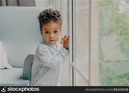 Portrait of adorable afro american curly haired baby boy in light blue colored shirt leaning on window glass in morning sunlight in living room at home while waiting for parents. Childhood concept. Portrait of cute mixed race curly haired baby boy waiting for parents near window