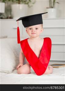 Portrait of adorable baby boy in graduation cap and red collar sitting on bed