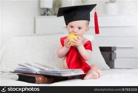 Portrait of adorable baby boy in graduation cap sitting in from of books and biting apple. Concept of baby growing wiser