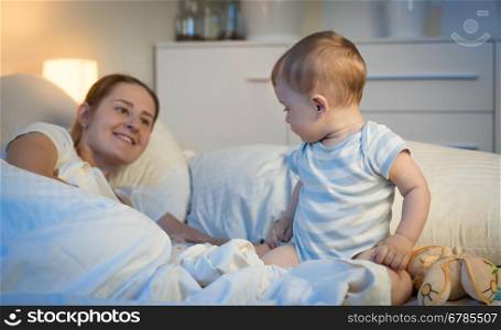 Portrait of adorable baby boy sitting on bed at night and looking at mother