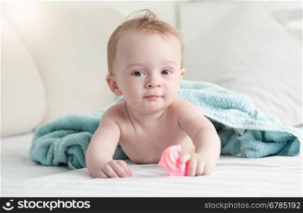 Portrait of adorable baby lying under blue towel on bed and holding plastic toy