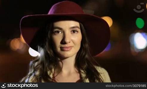 Portrait of adorable brunette female in fashionable bordo hat looking at camera and smiling at night city. Lovely long hair woman with deep brown eyes gazing at someone with enigmatic shy smile on colorful streetlights bokeh background.