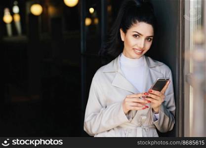 Portrait of adorable bruntte woman with dark eyes and eyebrows, pure healthy skin dressed in white clothes holding mobile phone in hands demonstrating her perfect manicure looking into camera