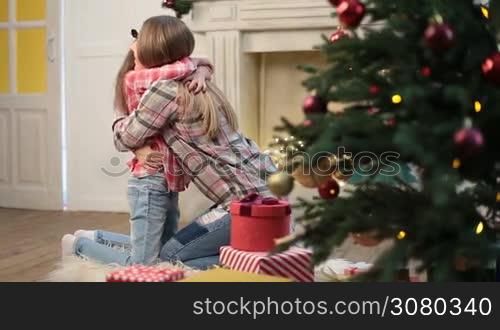 Portrait of adorable little daughter with long brown hair kissing and embracing attractive mother over christmas decorated room. Smiling cute girl hugging her mom during winter holidays as they both looking at camera and smiling. Dolly shot.