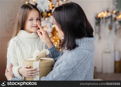 Portrait of adorable little kid and mother, touches her little nose, gives wrapped gift box, prepares little surpise for daughter, gives present near decorated New Year tree. Christmas and New Year