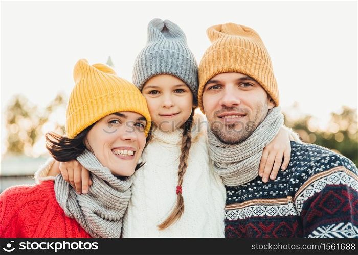 Portrait of adorable small girl wears knitted hat and sweater stands between parents, embrace them. Beautiful woman wears warm scarf and sweater enjoys free time with husband and little daughter