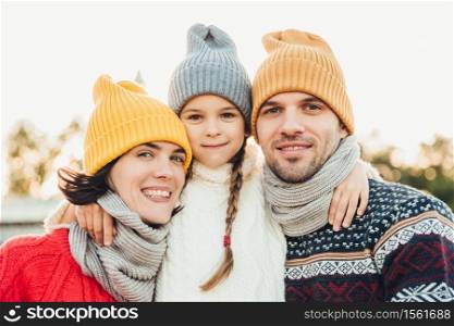 Portrait of adorable small girl wears knitted hat and sweater stands between parents, embrace them. Beautiful woman wears warm scarf and sweater enjoys free time with husband and little daughter