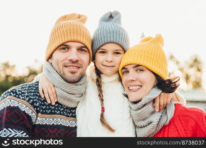 Portrait of adorable small girl wears knitted hat and sweater stands between parents, embrace them. Beautiful woman wears warm scarf and sweater enjoys free time with husband and little daughter