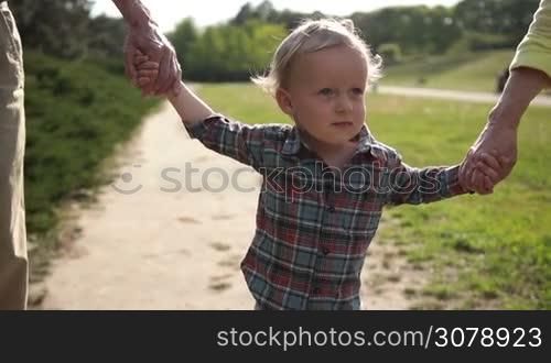Portrait of adorable smiling little toddler boy with blue eyes holding hands while walking with grandparents in park. Happy multi generation family having fun outside in spring nature. Slow motion. Steadicam stabilized shot.