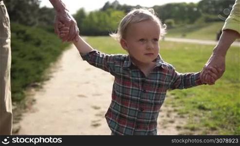 Portrait of adorable smiling little toddler boy with blue eyes holding hands while walking with grandparents in park. Happy multi generation family having fun outside in spring nature. Slow motion. Steadicam stabilized shot.