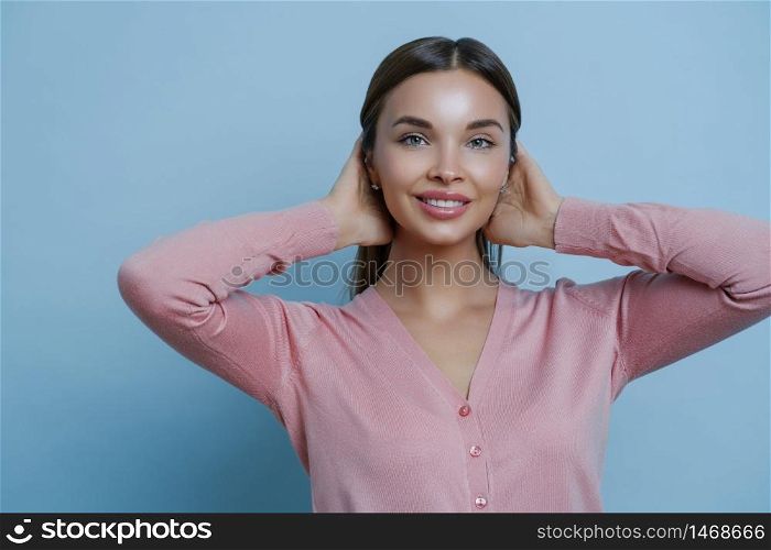 Portrait of adorable woman with perfect clean skin, toothy gentle smile, keeps hands on hair, wears rosy jumper, makes photo in studio, poses against blue background. Cosmetology and beauty concept
