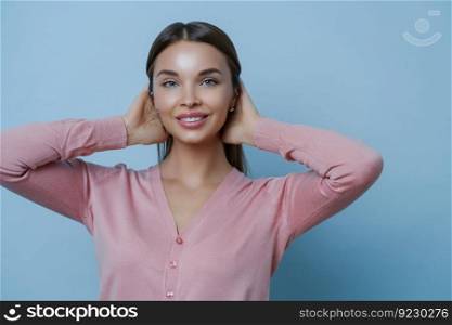 Portrait of adorable woman with perfect clean skin, toothy gentle smile, keeps hands on hair, wears rosy jumper, makes photo in studio, poses against blue background. Cosmetology and beauty concept