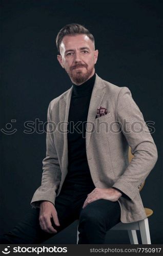Portrait of adult businessman wearing trendy suit and sitting in modern studio on stylish chair against the black background. Horizontal mockup. High quality photo. Portrait of adult businessman wearing trendy suit and sitting in modern studio on stylish chair against the black background