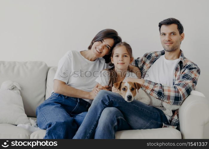 Portrait of affectionate family cuddle and sit together at couch in living room, change their home, have happy expressions. Father, mother, daughter and dog pose for making portrait, spend good time