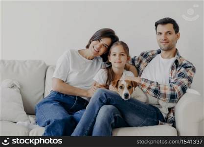 Portrait of affectionate family cuddle and sit together at couch in living room, change their home, have happy expressions. Father, mother, daughter and dog pose for making portrait, spend good time