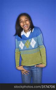 Portrait of African-American girl with hands in her pockets smiling standing against blue background.