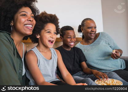 Portrait of African American grandmother, mother and children watching a movie and eating popcorn while sitting on sofa at home. Family and lifestyle concept.