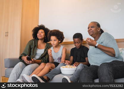 Portrait of African American grandmother, mother and children watching a movie and eating popcorn while sitting on sofa at home. Family and lifestyle concept.