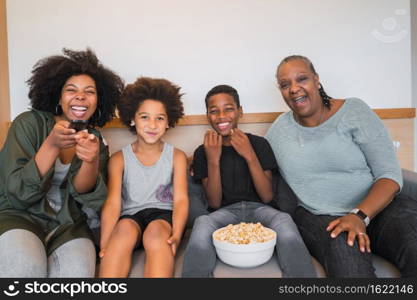 Portrait of African American grandmother, mother and children watching a movie and eating popcorn while sitting on sofa at home. Family and lifestyle concept.