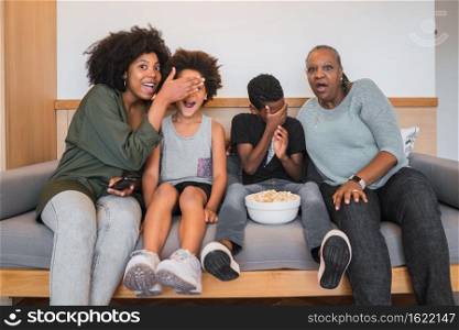 Portrait of African American grandmother, mother and children watching a scary movie and eating popcorn while sitting on sofa at home. Family and lifestyle concept.