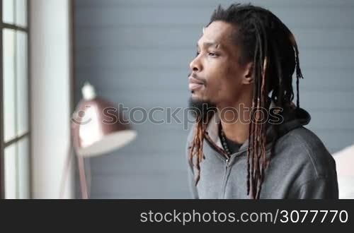 Portrait of african american hipster with dreadlocks looking at the camera and smiling in domestic interior. Closeup of handsome stylish man with toothy smile gazing at someone in slow motion.