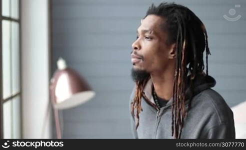 Portrait of african american hipster with dreadlocks looking at the camera and smiling in domestic interior. Closeup of handsome stylish man with toothy smile gazing at someone in slow motion.