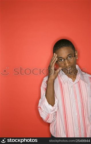 Portrait of African-American teen boy with glasses and hand on forehead against red background.