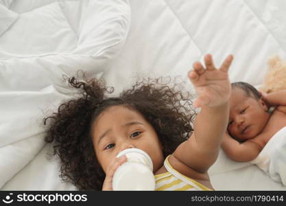 Portrait of African little girl is lying in bed and sucking a milk bottle with her newborn baby boy and looking at camera. New siblings relationship in bedroom at home with love