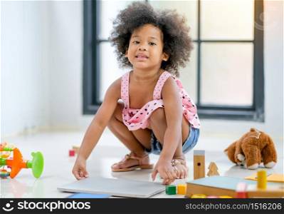 Portrait of African little girl sit in squat position and smile among several toys and also hold thin laptop in the living room with day light.