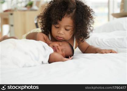Portrait of African newborn baby sleeping on a white bed. His little sister kissing infant boy with love. New siblings relationship in bedroom at home