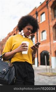 Portrait of Afro American latin woman using her mobile phone while holding a cup of coffee outdoors in the street. Urban and communication concept.