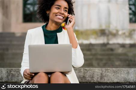 Portrait of afro business woman talking on phone and using her laptop while sitting on stairs outdoors. Business concept.