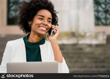 Portrait of afro business woman talking on phone and using her laptop while sitting on stairs outdoors. Business concept.