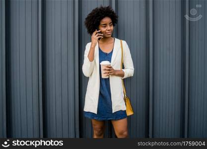 Portrait of afro business woman talking on the phone and holding a cup of coffee while standing outdoors at the street. Business and urban concept.