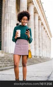 Portrait of afro business woman using her mobile phone and holding a cup of coffee while walking outdoors at the street. Business and urban concept.. Business woman using her mobile phone outdoors.
