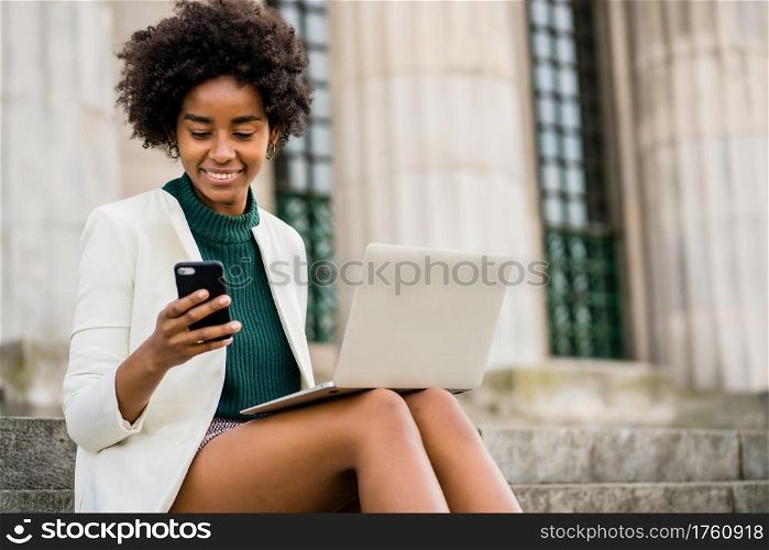 Portrait of afro business woman using her mobile phone and laptop while sitting on stairs outdoors. Business concept.