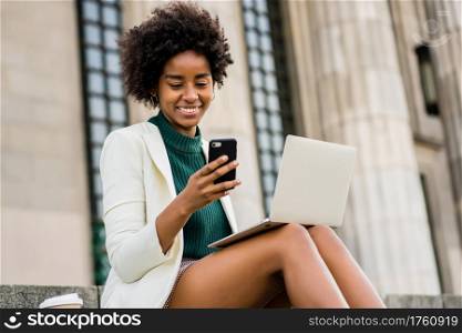 Portrait of afro business woman using her mobile phone and laptop while sitting on stairs outdoors. Business concept.