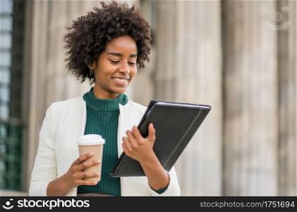Portrait of afro businesswoman holding a cup of coffee and looking at clipboard while standing outdoors at the street. Business and urban concept.