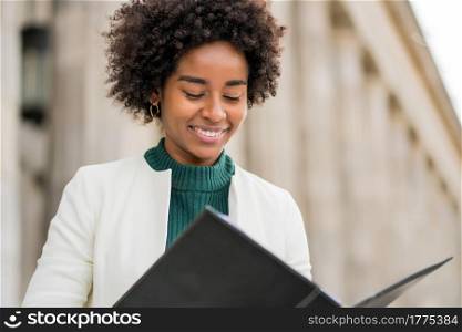 Portrait of afro businesswoman holding and looking at clipboard while standing outdoors at the street. Business and urban concept.