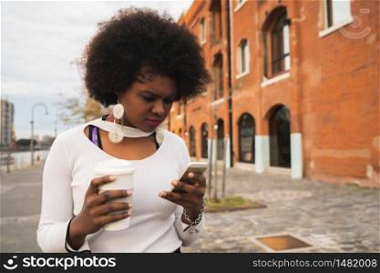 Portrait of Afro latin woman using her mobile phone while holding a cup of coffee outdoors in the street. Urban and communication concept.