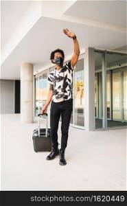 Portrait of afro tourist man carrying suitcase and raising his hand to hail a taxi while walking outdoors on the street. Tourism concept.