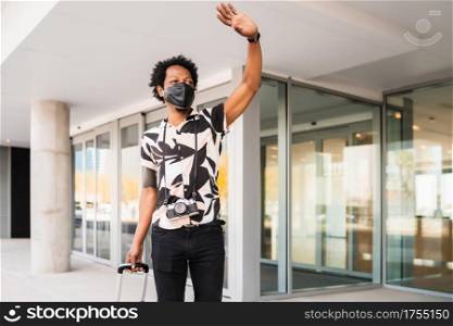 Portrait of afro tourist man carrying suitcase and raising his hand to hail a taxi while walking outdoors on the street. Tourism concept.