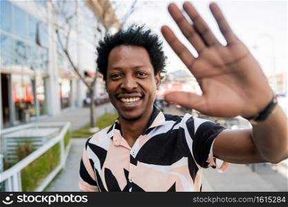 Portrait of afro tourist man saying hi with hand while standing outdoors on the street. New normal life concept.