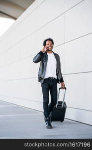 Portrait of afro tourist man talking on the phone and carrying suitcase while walking outdoors on the street. Tourism concept.