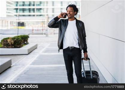 Portrait of afro tourist man talking on the phone and carrying suitcase while walking outdoors on the street. Tourism concept.