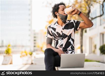 Portrait of afro tourist man using his laptop and drinking a cup of coffee while sitting outdoors. Tourism concept.
